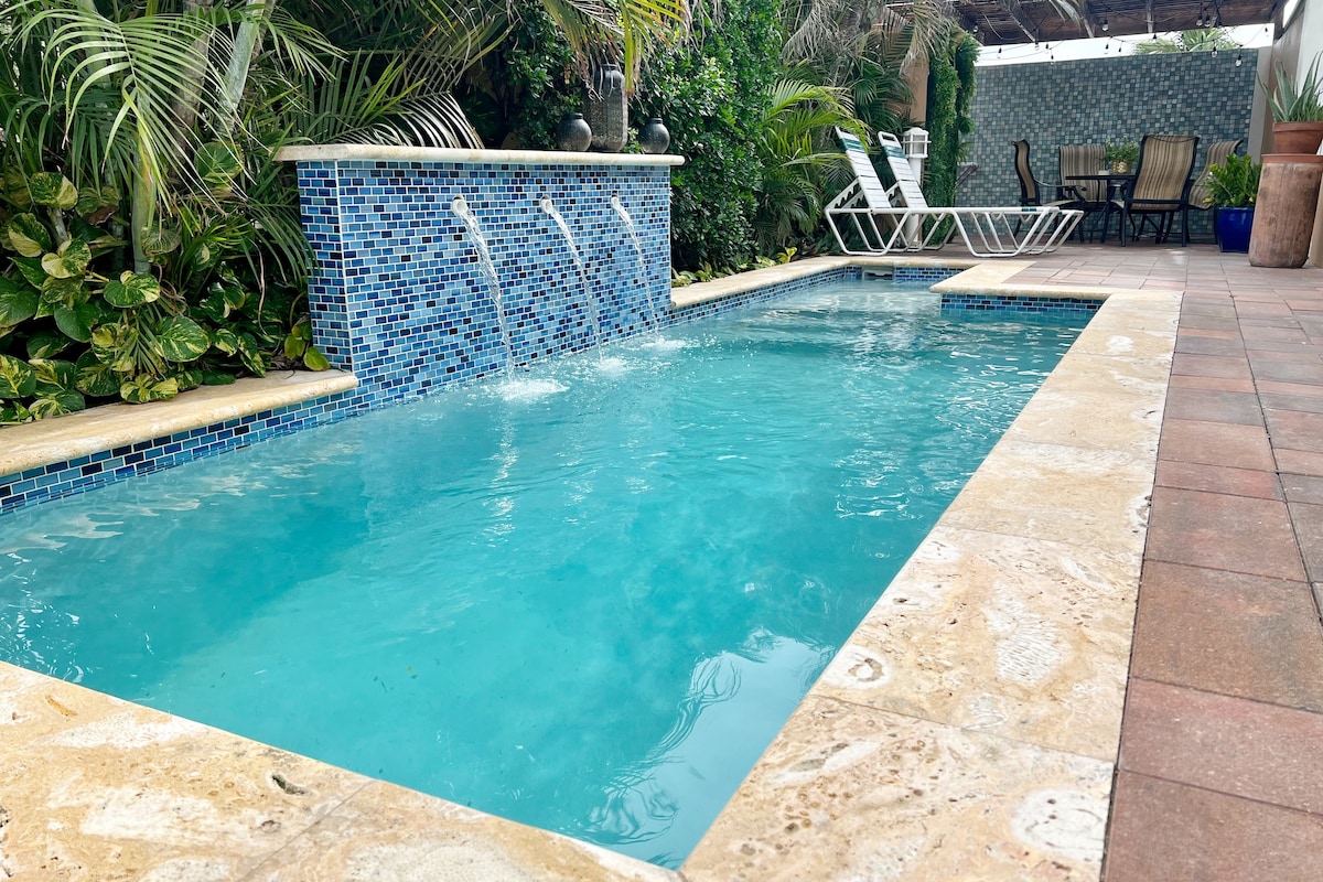 A private pool is framed by vibrant greenery and a mosaic tile wall. Water features create a soothing atmosphere. Lounge chairs are positioned nearby, inviting relaxation in the tranquil setting, while an outdoor dining area is visible in the background.