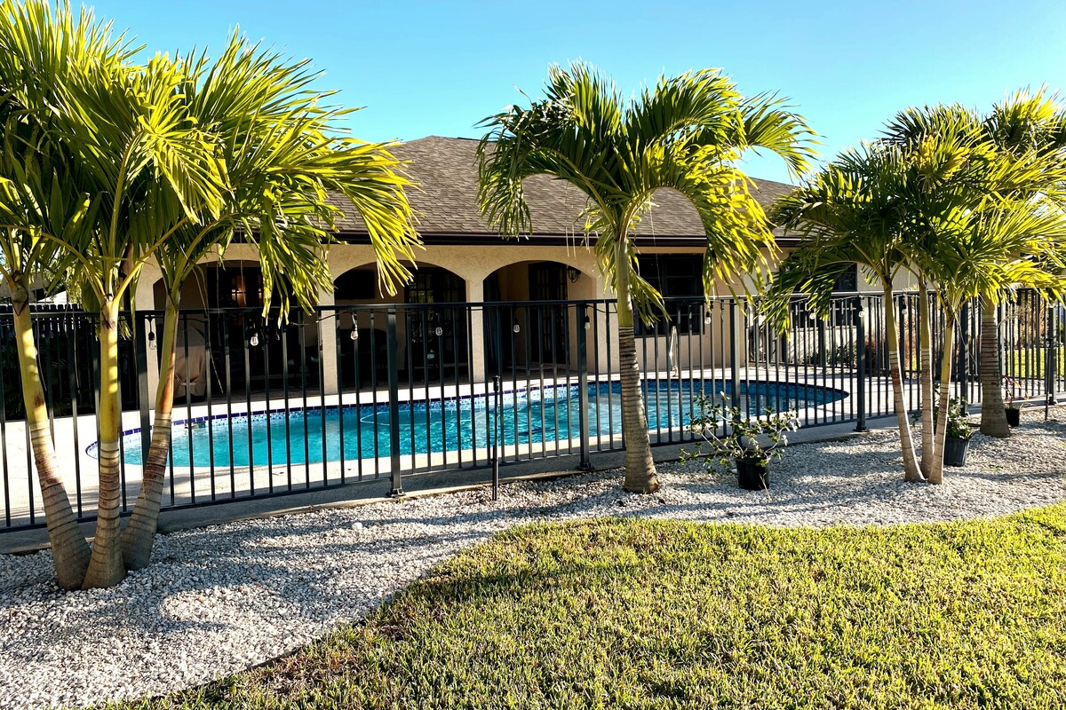 A backyard scene features a fenced pool surrounded by lush palm trees. Gravel landscaping complements the green grass, creating a tranquil outdoor space. The house is visible in the background, showcasing large windows that open toward the pool area.