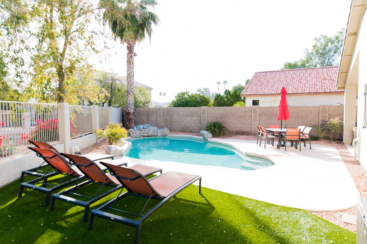A spacious backyard is presented, featuring a private pool with a waterfall feature and surrounded by lush artificial turf. Four loungers are positioned beside the pool, and a dining set is shown under a red umbrella, complemented by desert landscaping along the perimeter.