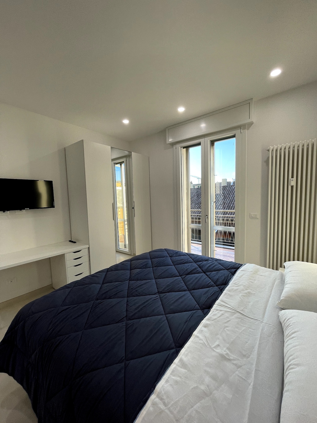 A spacious bedroom features a large bed covered with a navy blue quilt. Natural light enters through large double windows, which offer a view of the exterior balcony. A flat-screen television is mounted on the wall opposite a modern desk with storage.