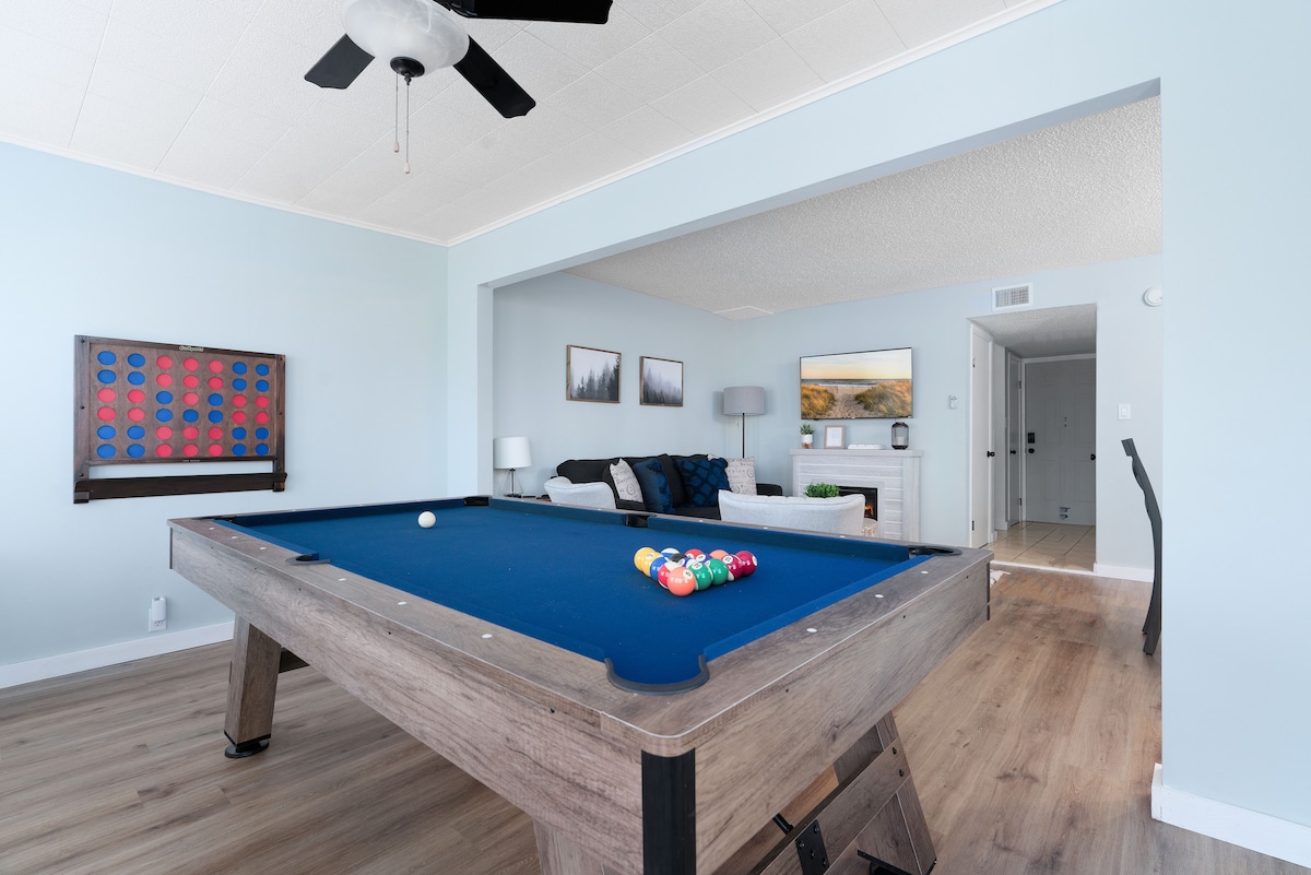 A pool table with a blue felt surface is positioned in a spacious living area. Colorful pool balls are neatly arranged on the table. Light wood flooring complements the light blue walls, while a ceiling fan adds comfort to the space.
