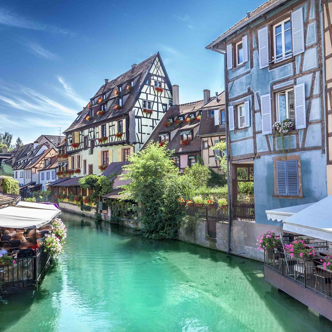 Charming half-timbered houses line a tranquil canal, reflecting traditional Alsatian architecture. Lush greenery and vibrant flowers enhance the scene, while calm waters display a soothing turquoise hue. A bright blue sky provides a serene backdrop, inviting exploration of the picturesque surroundings.