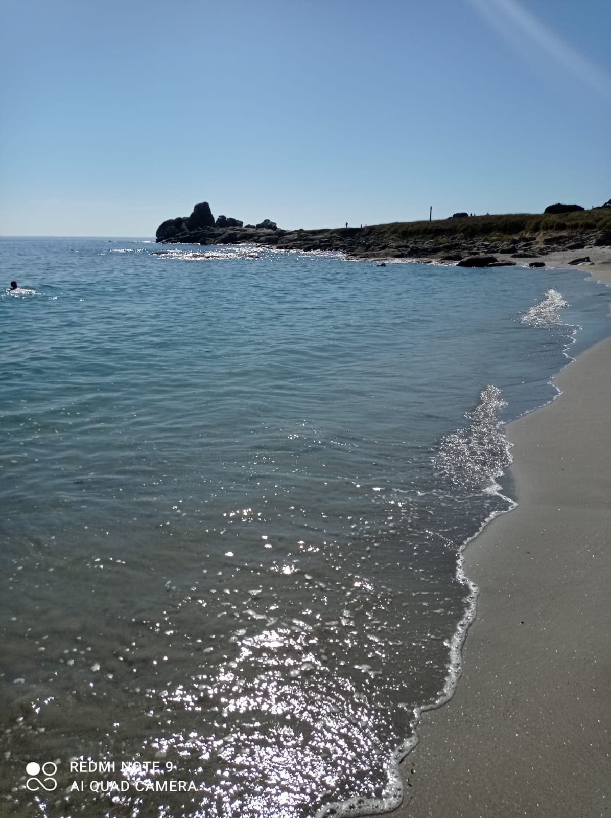 A serene beach scene is featured, with gentle waves lapping against a sandy shore. Sunlight sparkles on the water, creating a calm atmosphere. In the distance, rocky formations are visible against a clear blue sky, providing a natural backdrop.