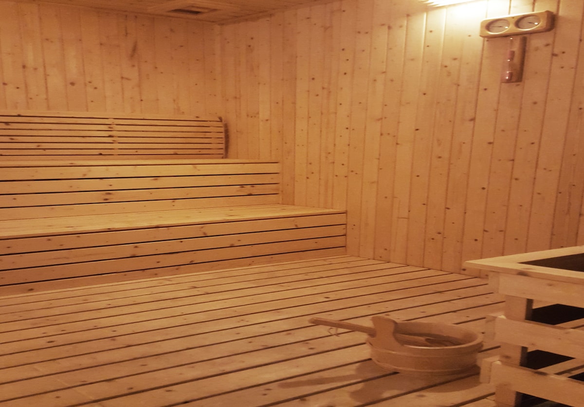 The sauna features warm wooden paneling throughout, creating a soothing environment. Benches are arranged in tiered levels for seating. A wooden bucket and ladle are positioned on the floor, complementing the tranquil atmosphere.