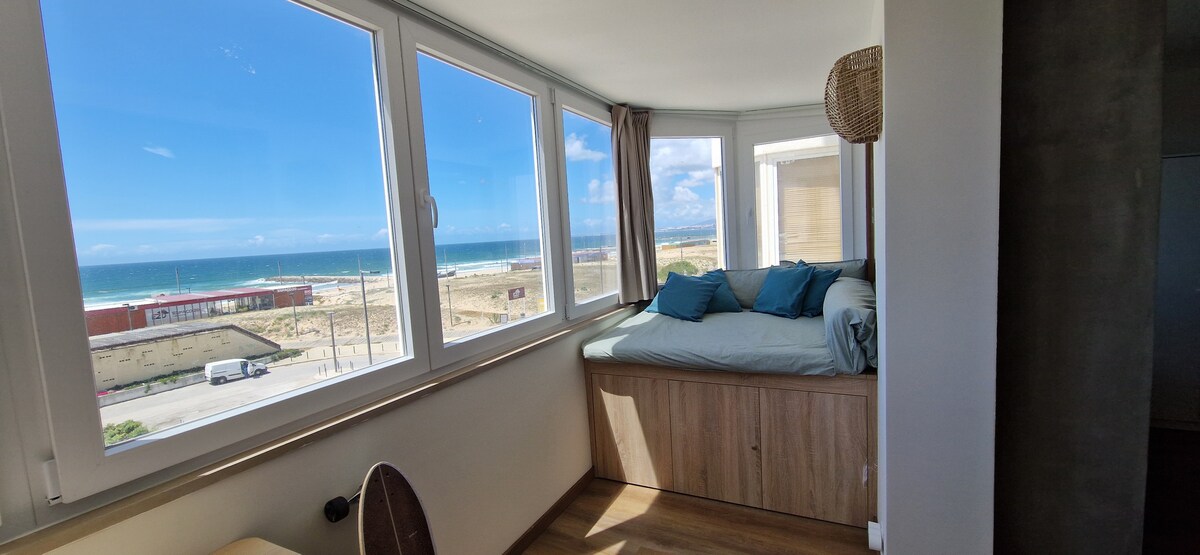 Large windows frame a scenic view of the Atlantic Ocean, floods the cozy nook with natural light. A spacious bed adorned with blue cushions is positioned against the wall in a corner, blending comfort with a serene outlook. Coastal landscapes and sandy shores are visible beyond.