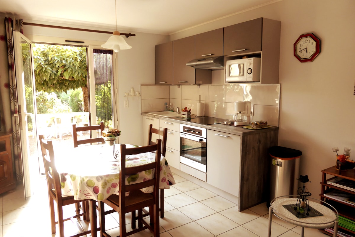 A bright kitchen area features a dining table with wooden chairs, surrounded by light-colored cabinetry and a modern stove. Large glass doors open to an outdoor space, allowing natural light to enhance the room's airy feel. A clock and a small trash can are visible.
