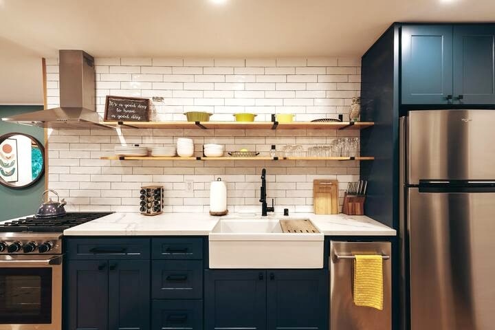 A modern kitchen showcases stainless steel appliances and quartz countertops. Open shelving displays colorful dishes and glassware above a spacious farmhouse sink. A gas range is positioned next to a sleek refrigerator, complementing the vintage-style subway tile backsplash and navy blue shaker cabinets.