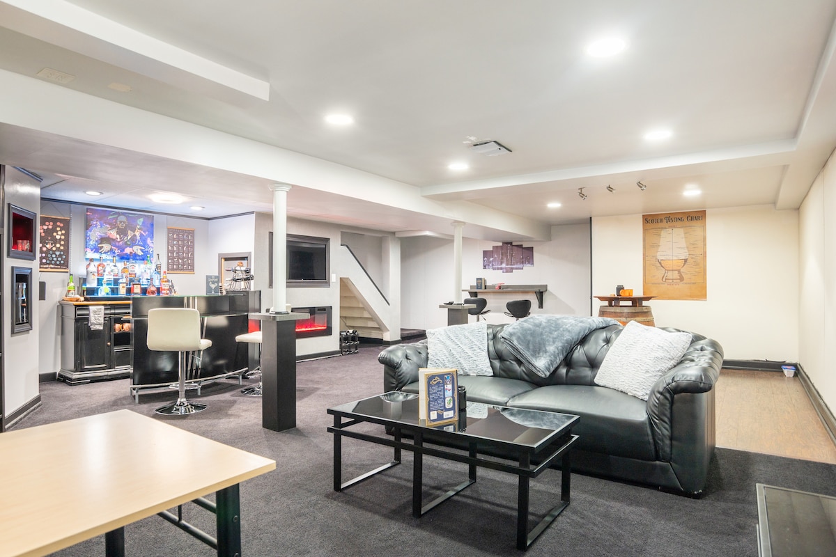 A spacious entertainment area is presented, featuring a comfortable leather sofa arranged around a glass coffee table. A full wet bar with various glassware and a smart TV are visible in the background. The area is well-lit, with modern decor and an inviting ambiance.