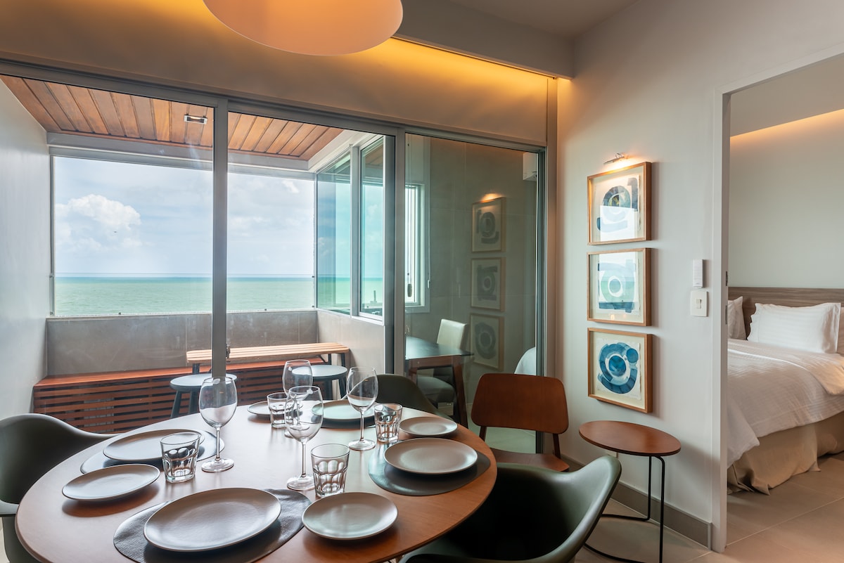 A dining area is equipped with a round wooden table set with plates and glassware, complemented by chairs. Floor-to-ceiling windows provide views of the ocean, while soft natural light enhances the serene atmosphere. In the background, a cozy sleeping area is partially visible.