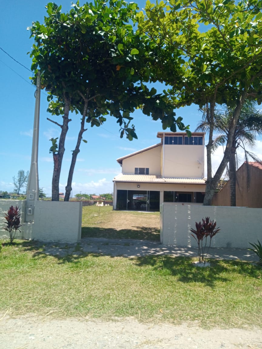 The exterior of a spacious two-story home is shown, framed by tall trees and a clear blue sky. A wide lawn extends in front of the entrance, leading to a welcoming gateway, while the house features large windows that offer a bright and open appearance.
