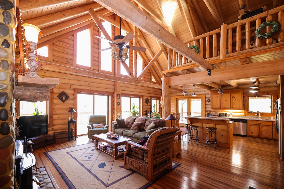 The spacious living area features a high, vaulted ceiling supported by exposed wooden beams. Large windows allow ample natural light, highlighting a comfortable seating arrangement and a coffee table on a patterned area rug. A dining area and open kitchen are visible in the background.