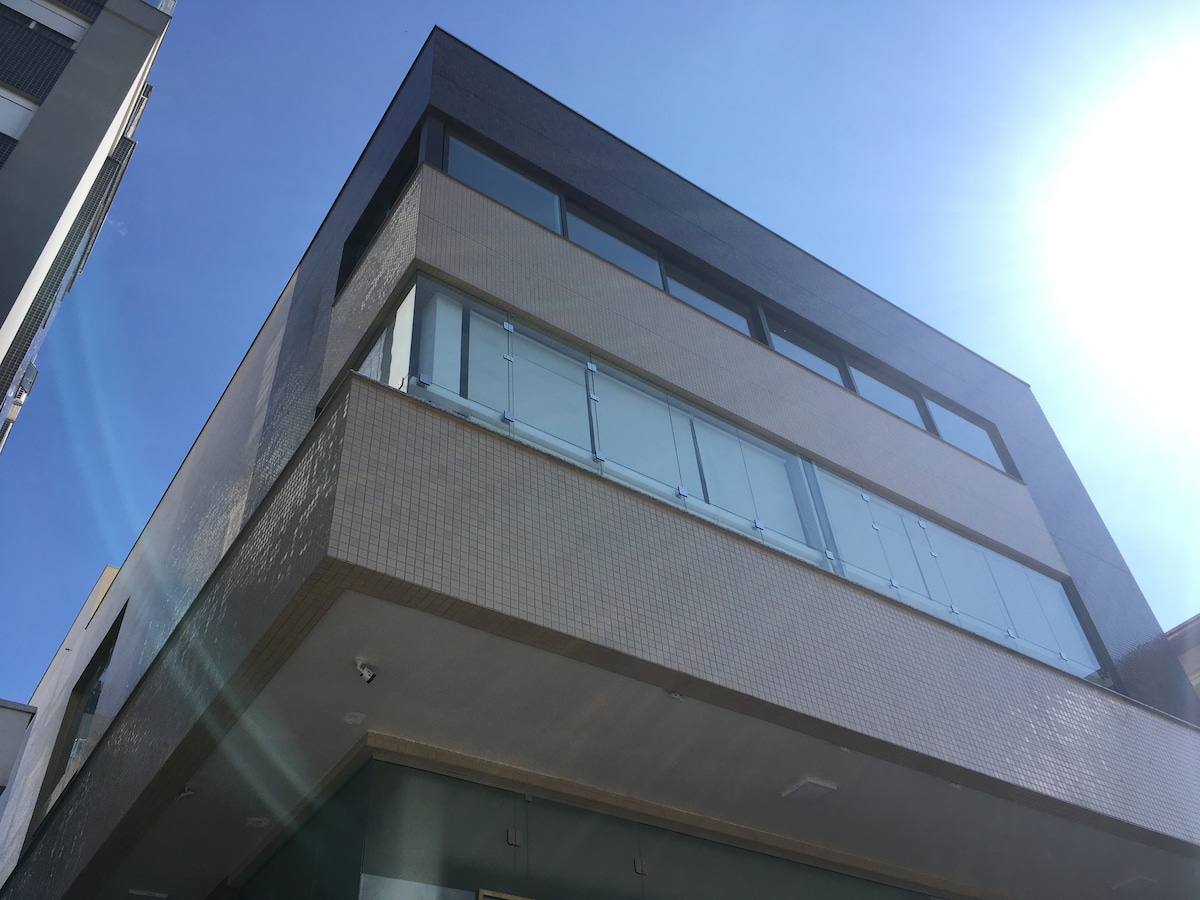 The exterior of a modern building is captured from a low angle, highlighting a sleek design and large windows on the upper floors. A balcony with glass railings is visible, allowing for natural light and open views. Clear blue skies provide a bright backdrop.