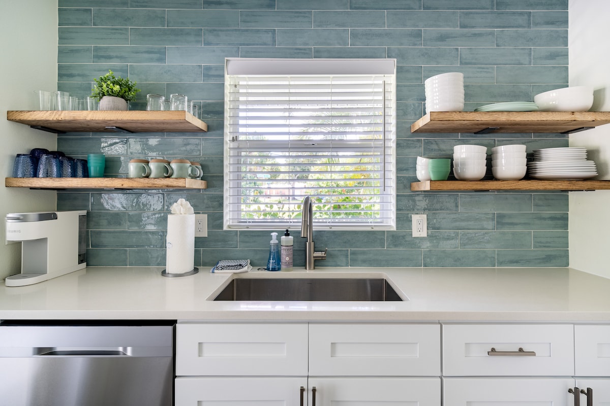 A modern kitchen area is presented with light wood shelves displaying various dishware and glassware. A stainless-steel sink is centered beneath a bright window, allowing natural light to illuminate the space. Neutral countertops complement the calm blue tones of the tiled backsplash.
