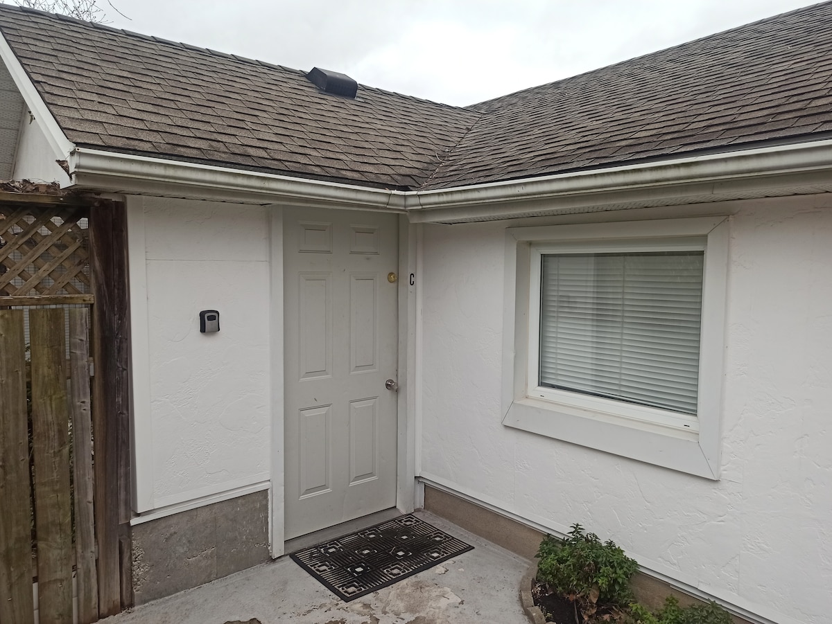 The exterior of the guesthouse is presented, featuring a neutral-colored facade with a single door and a window. A welcome mat lies at the entrance, while a small planter sits nearby. The roof is covered with shingles, and there is a wooden fence in the background.