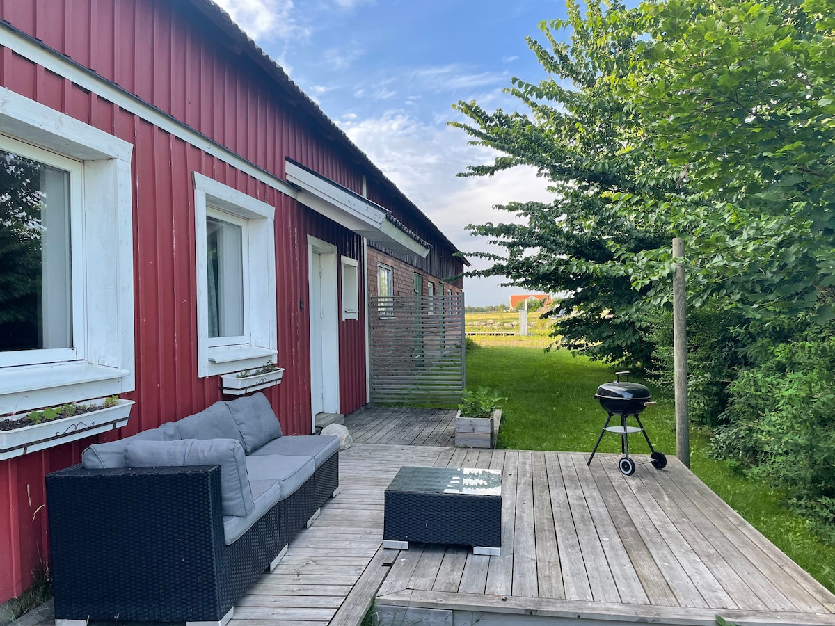 An outdoor patio area is available, featuring a seating arrangement with a dark wicker sofa and a glass-top coffee table. A small charcoal grill is positioned on a wooden deck, surrounded by greenery and fields in the background.