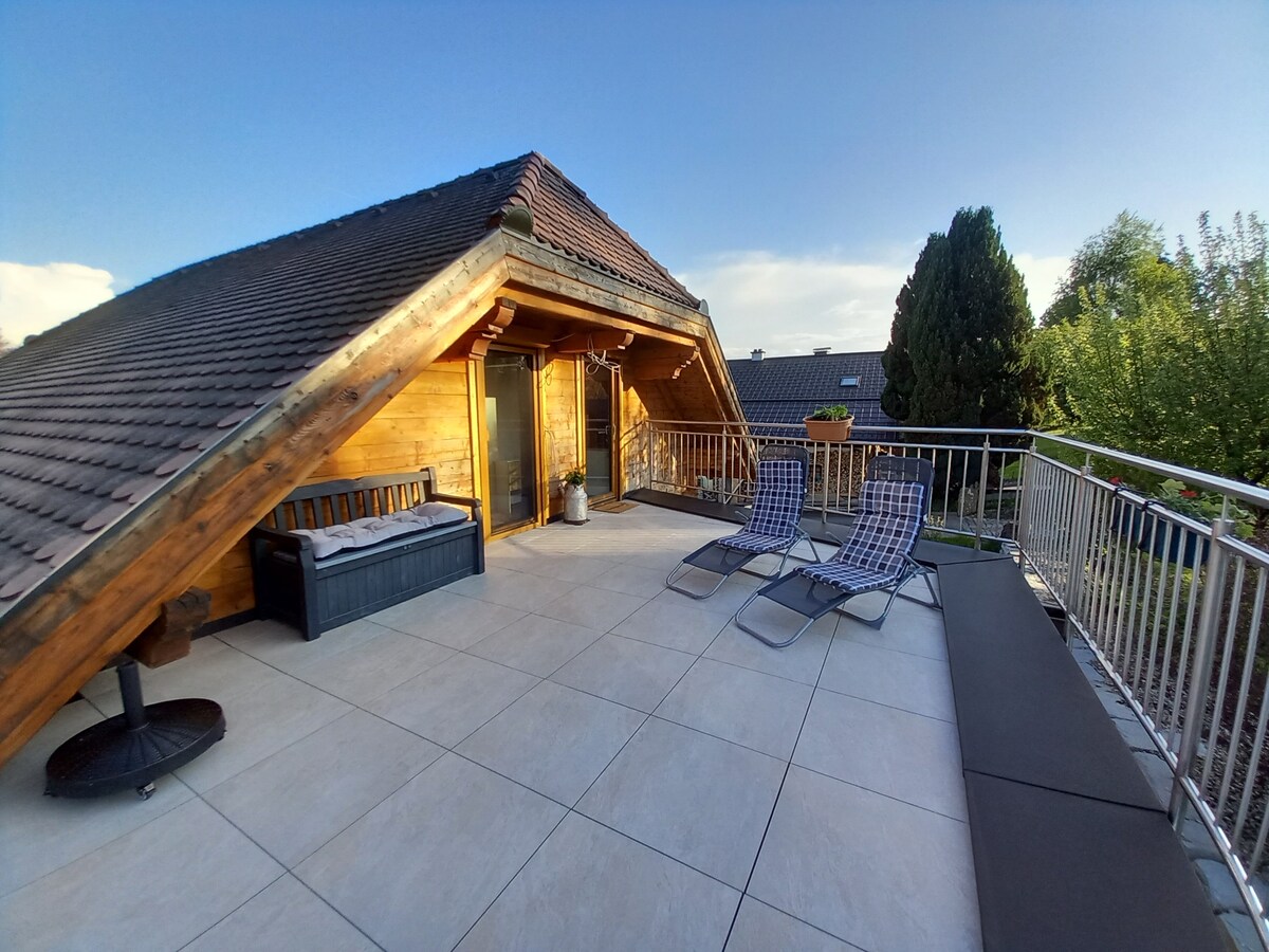 A spacious terrace is featured, surrounded by wooden walls and a sloped roof. Two lounge chairs are positioned near a sunbed, with a stylish seating area visible. The area is illuminated by natural light, enhancing the inviting outdoor space.