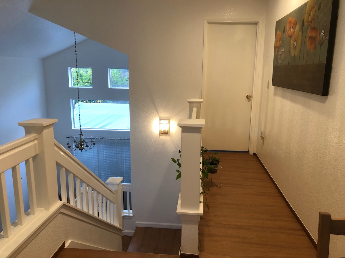 A bright hallway is visible with new flooring and white walls. Natural light streams through large windows at the end, illuminating a decorative wall art featuring flowers. A plant is placed by the staircase railing, contributing to the inviting ambiance.