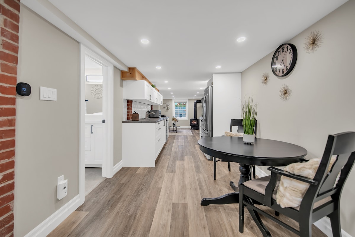 A spacious kitchen area with modern cabinetry and sleek countertops is seen leading into a dining space. A round table with two chairs in a minimalist style is situated to the right, while a clock adorns the wall opposite.