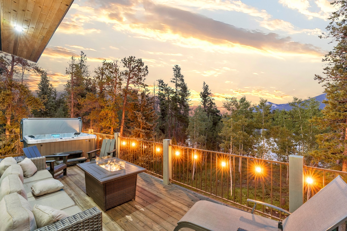 A wooden deck features a spacious hot tub overlooking a serene landscape with trees and distant mountains. Comfortable outdoor seating surrounds a gas fire pit, while string lights add a soft glow to the setting, enhancing the tranquil ambiance at sunset.