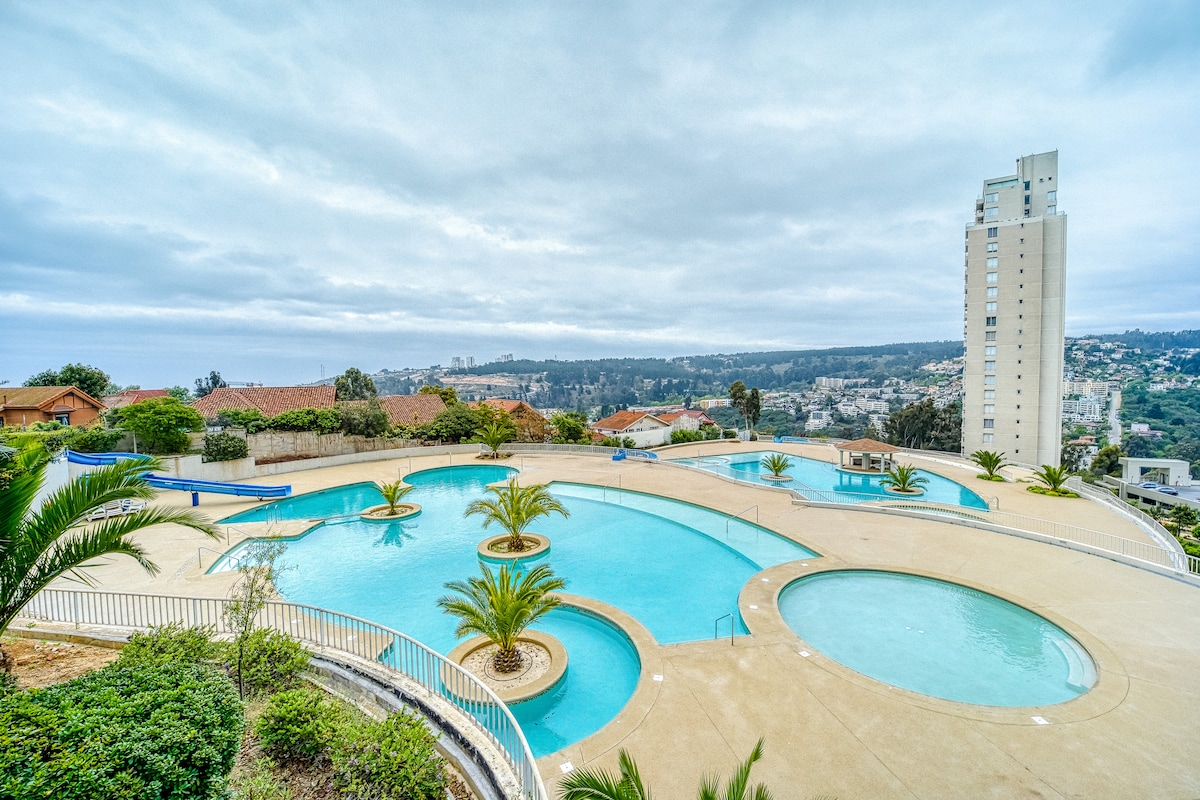 An expansive pool area is surrounded by landscaped gardens and palm trees. Multiple pools of varying shapes are visible, along with lounge spaces. A tall building stands in the background, complementing the scenic view of the hills and city beyond.