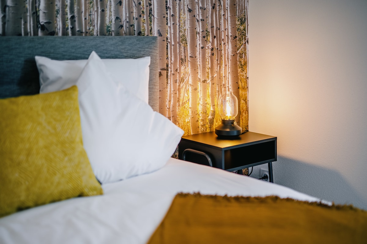 A cozy sleeping area features a spacious bed adorned with soft white pillows and a yellow decorative throw. A stylish bedside table holds an illuminated lamp, casting a warm glow against the tree-patterned wallpaper in the background.