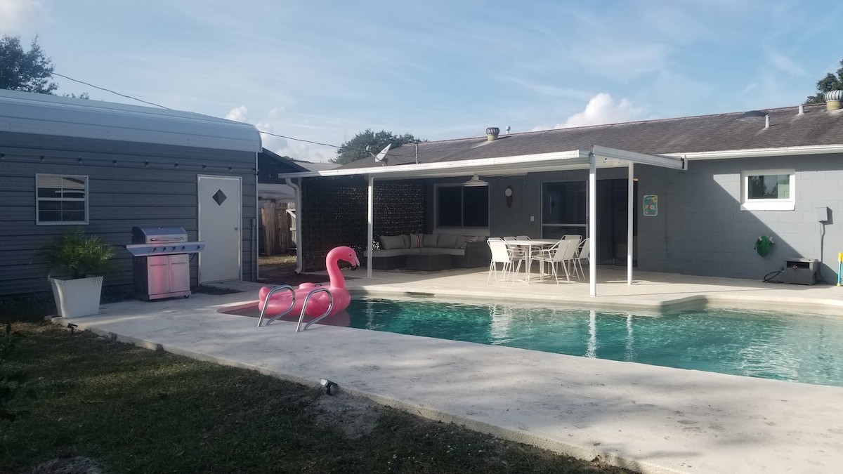 An outdoor pool area is highlighted by a clear swimming pool and two pink flamingo floats. A covered patio with dining furniture overlooks the pool, providing spaces for relaxation. A grill is positioned nearby under an overhang, surrounded by a lawn area and a shed.