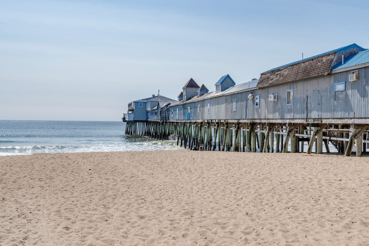 A wooden pier extends over the sandy beach, supported by sturdy stilts. The calm ocean stretches into the distance, with gentle waves lapping at the shore. A collection of beach structures can be seen along the pier, featuring a variety of architectural elements.