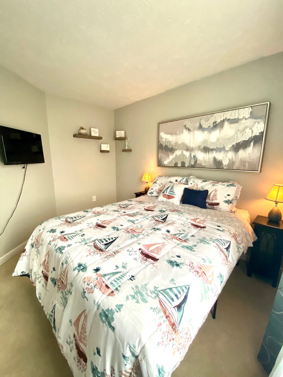 The bedroom features a cozy queen-size bed adorned with a patterned duvet cover depicting sailboats. Two bedside tables hold lamps, providing soft lighting. A mounted television is positioned on the wall opposite the bed, and decorative shelves enhance the room's ambiance.