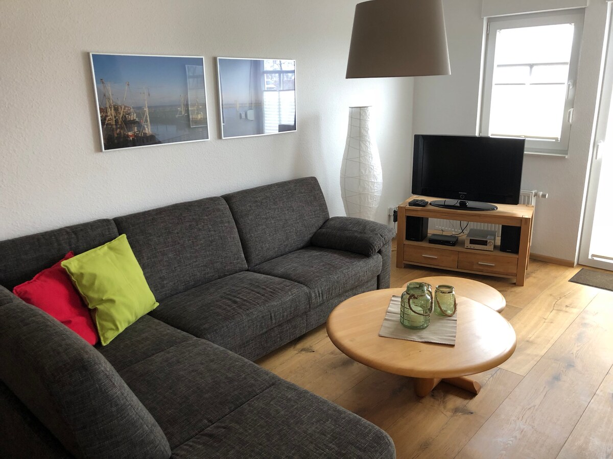 A spacious living area is furnished with a gray sectional sofa, complemented by colorful pillows. A round wooden coffee table sits in the center, accompanied by decorative glass items. A flat-screen TV is positioned on a wooden media unit, with natural light entering through a nearby window.