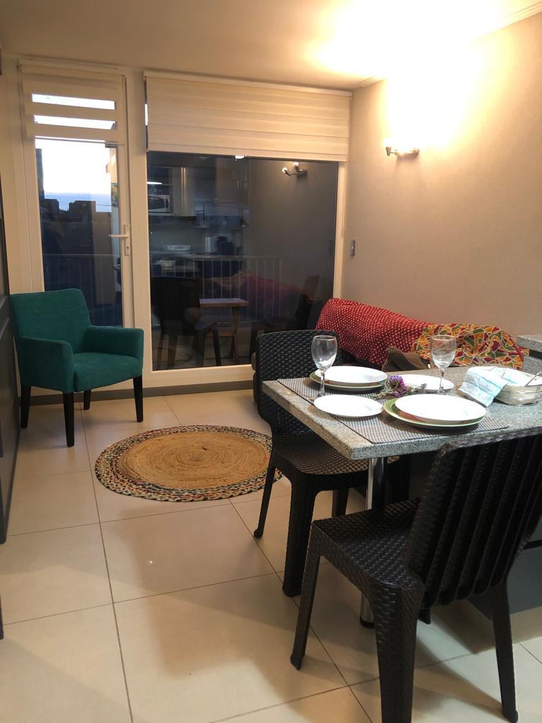 The dining area is furnished with a table set for two, accompanied by four black wicker chairs. A turquoise armchair is positioned nearby. Natural light filters through large windows, revealing an outdoor view and a vibrant throw covering the back of the chair.