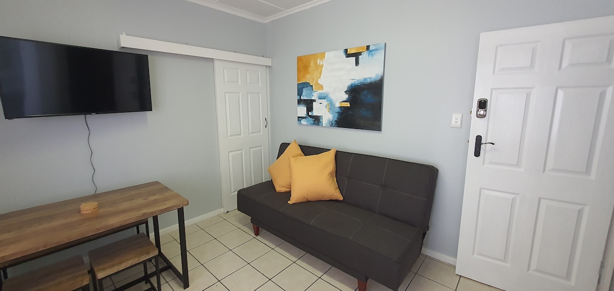 A modern living area features a sleek sleeper couch adorned with yellow pillows. A wooden table with stools is positioned nearby. A flat-screen television is mounted on the wall, and a colorful abstract painting complements the light-colored walls.