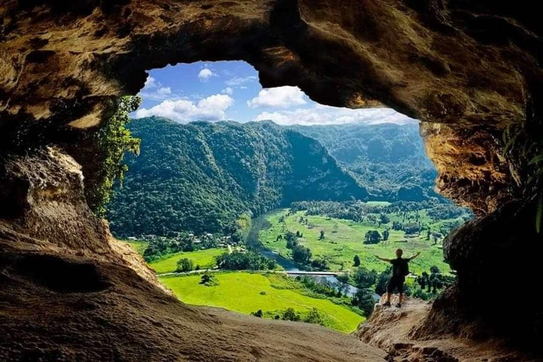 A wide view of a lush green valley framed by a cave opening is displayed. Rolling hills and a river can be seen in the distance under a clear blue sky, providing a natural backdrop. A person stands at the edge of the cave, enhancing the sense of scale.