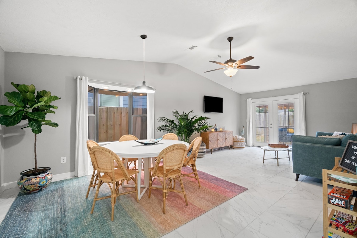 A light-filled dining area features a white table surrounded by six rattan chairs. A colorful area rug adds warmth to the spacious tile floor. Nearby, a ceiling fan circulates air, and a cozy seating area is positioned in an inviting corner of the room.