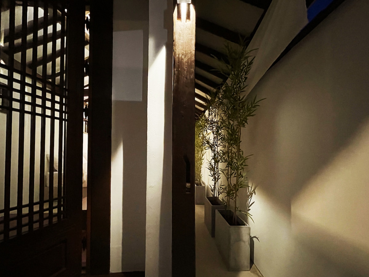 A tranquil corridor is bordered by a white wall and illuminated by soft lighting. Bamboo plants in large gray planters provide a fresh touch to the space, while traditional wooden beams accentuate the serene ambiance of the entrance.