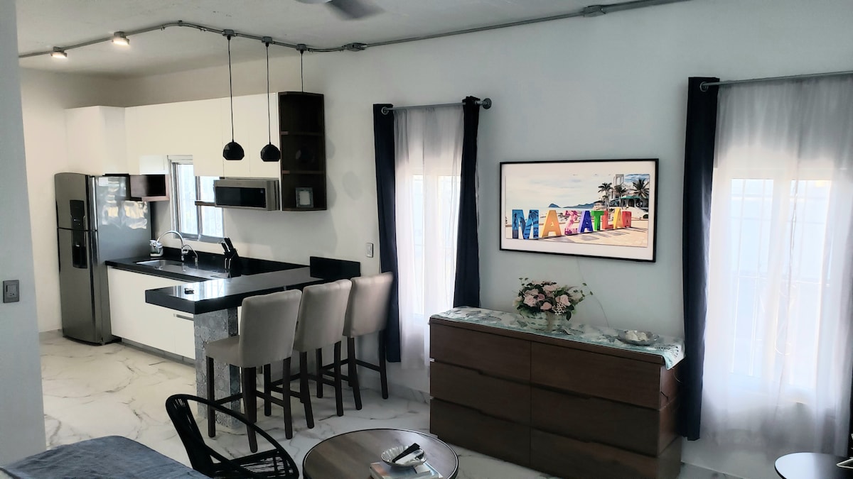 A modern studio apartment is displayed, featuring a sleek kitchen with black cabinetry and a dark countertop. Seating for four is provided at a minimalist dining area. A colorful artwork titled 'Mazatlán' is mounted on a wall, complemented by light curtains that soften the natural light.