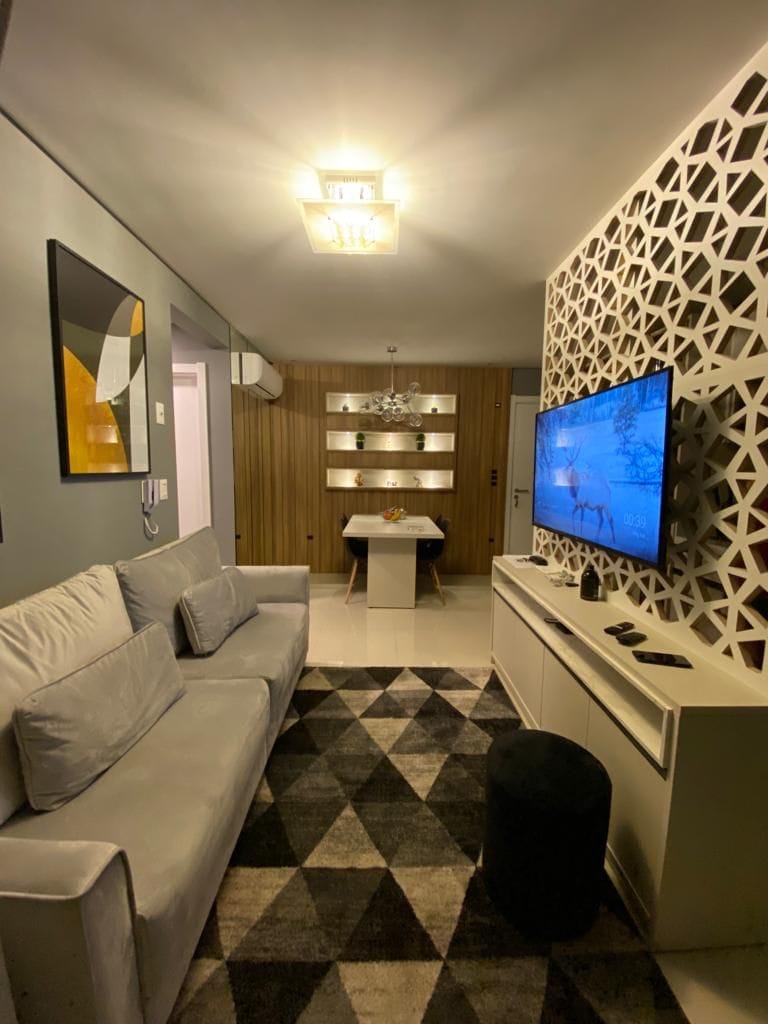 A modern living area is displayed, featuring a light gray sofa and a geometric accent wall. A large television is mounted on a white entertainment unit, with decorative items above. A dining table with white legs and chairs is visible, complemented by a patterned rug.