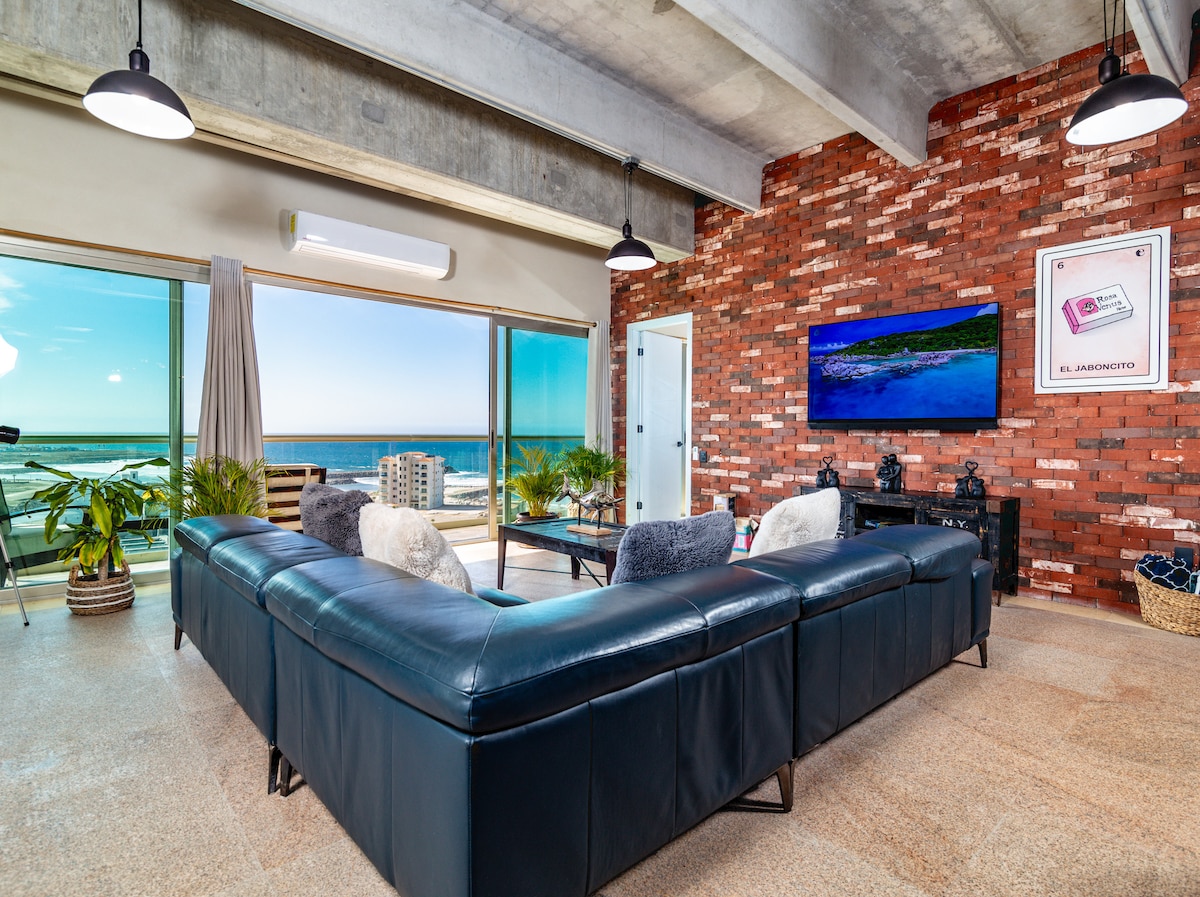 A spacious living area features a large sectional sofa overlooking panoramic ocean views through expansive glass doors. Natural light illuminates the room, highlighting the textured brick wall and modern decor. A television is mounted on the wall, and tropical plants add a touch of greenery.