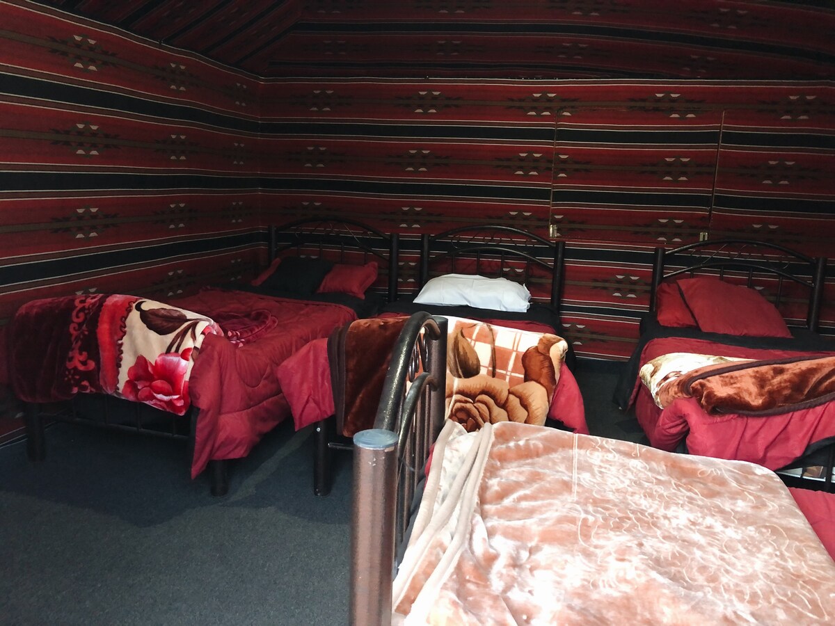 A cozy sleeping area features multiple beds with warm blankets in shades of red and brown. The walls are adorned with patterned fabric, adding to the room's distinctive ambiance. Natural light is present, creating a comfortable and inviting space.