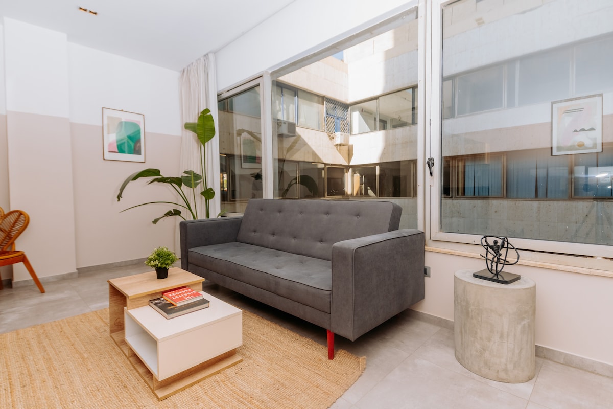 A modern living area is presented with a gray sofa facing large glass windows that bring in natural light. A round coffee table sits on a textured area rug, complemented by a small planter and an abstract decor piece on the side.