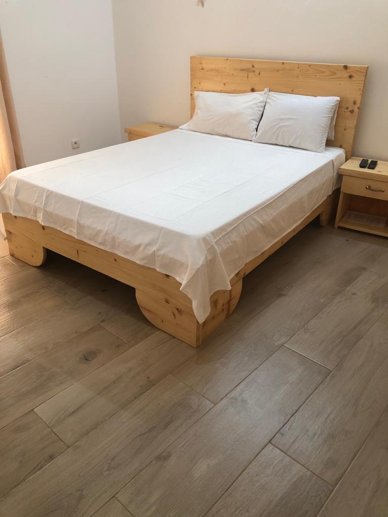 A spacious bed with a light-colored wooden frame is positioned against a plain wall. Two white pillows rest atop a neatly tucked white sheet. Simple nightstands are located on either side of the bed, enhancing the room's clean and minimalist aesthetic.
