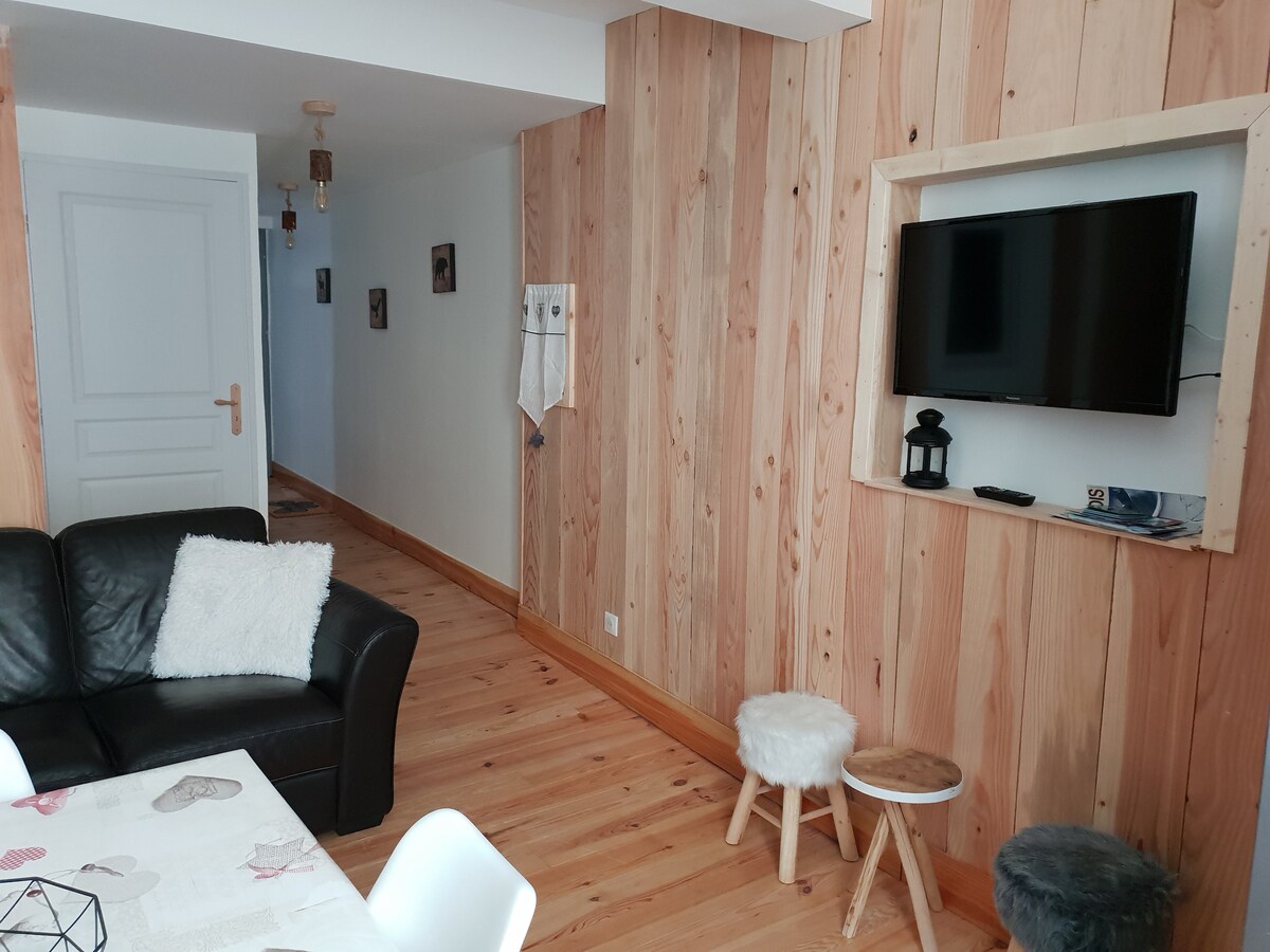 A living area is showcased with natural wood paneling, enhancing a modern yet rustic feel. A black leather sofa is positioned beside a light-colored table. A wall-mounted television is framed in wood, with a small lantern and magazines nearby. The floor features warm wooden tones.