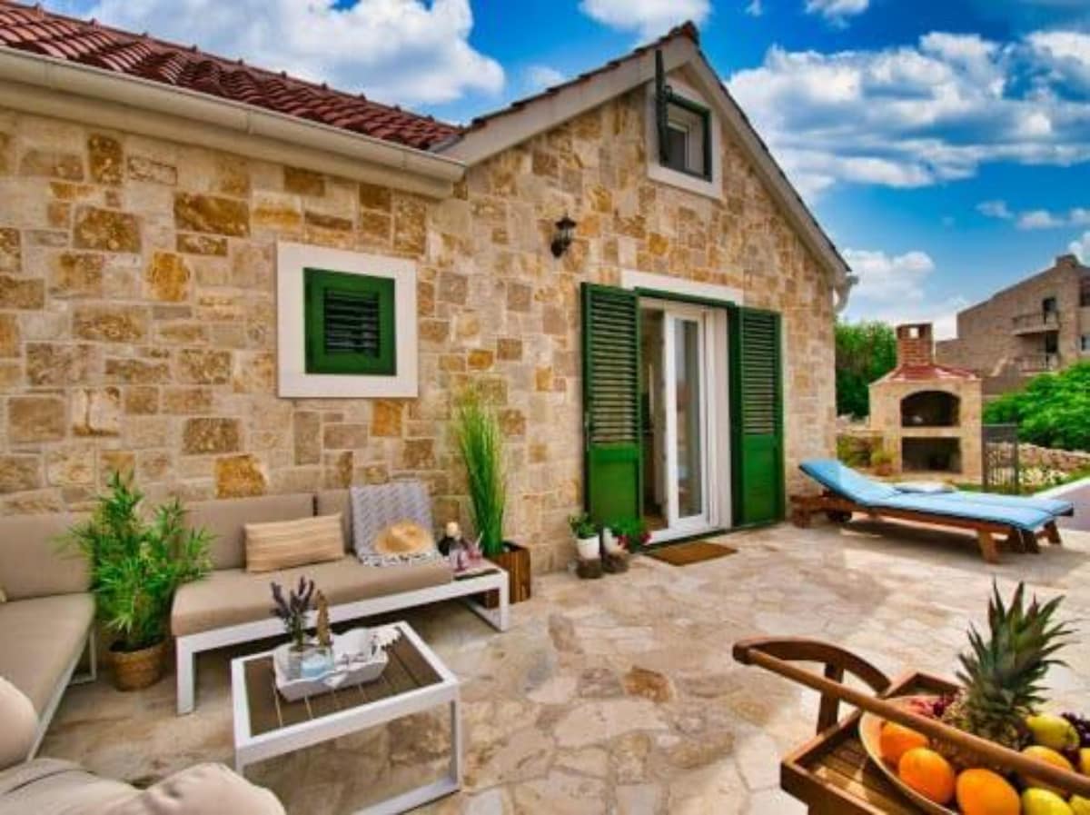 A stone patio is adorned with comfortable seating and decorative plants. A barbecue area and a sun lounger are presented, inviting relaxation. Bright green shutters frame the patio door, while a wooden cart displays fresh fruit, enhancing the outdoor living space.