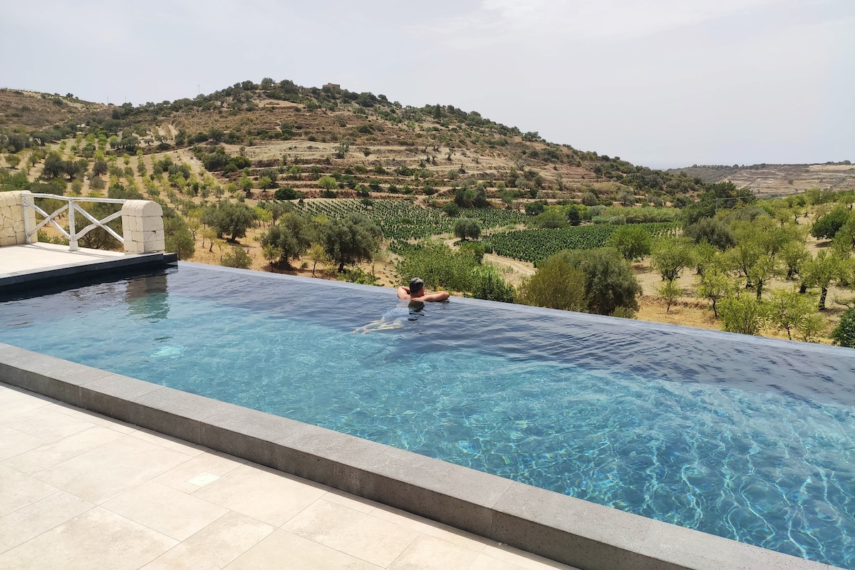 An infinity pool stretches along a terrace, offering sweeping views of the surrounding hills dotted with olive and almond trees. A swimmer is seen enjoying the tranquil water under a clear sky, blending with the serene landscape.