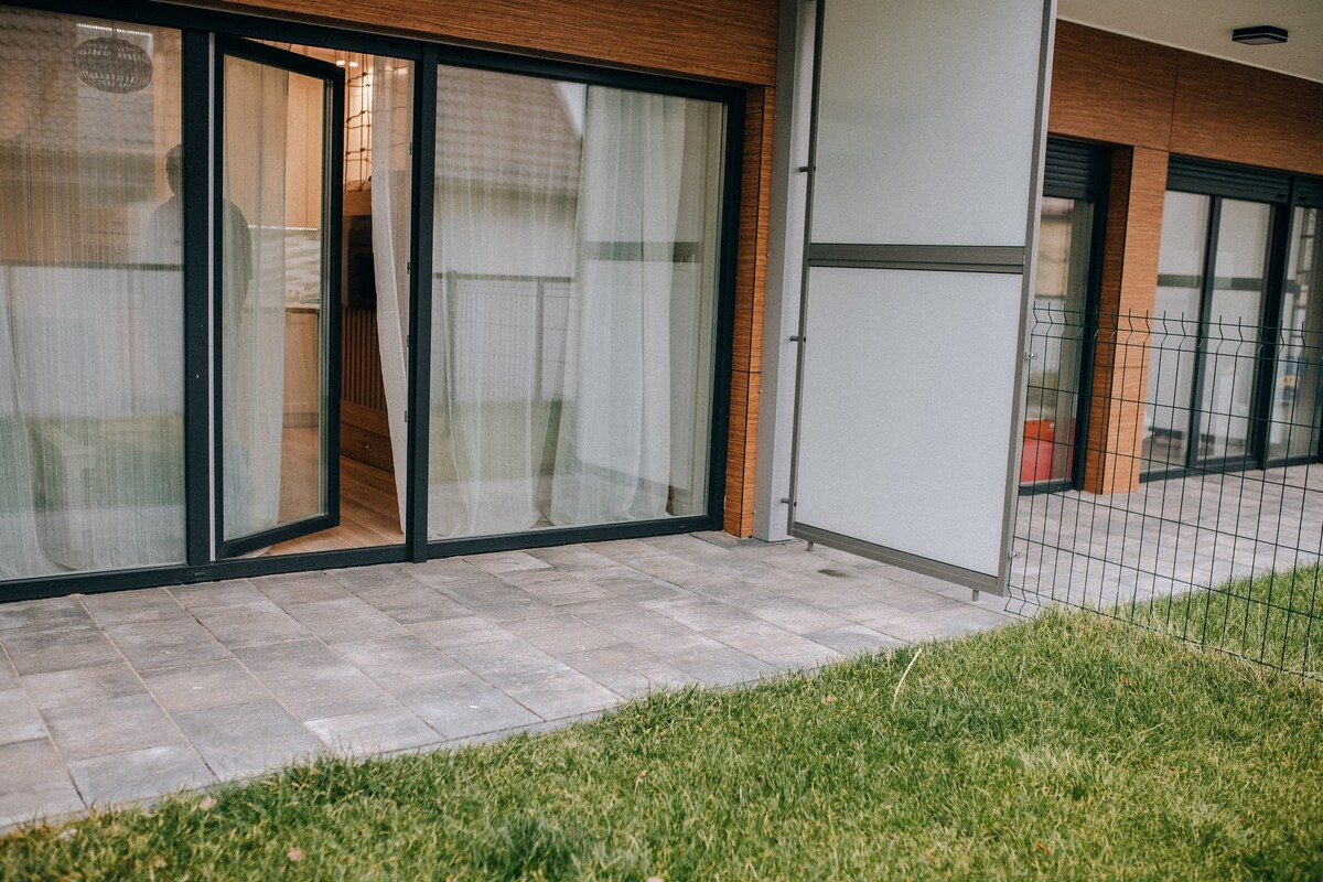 A patio area is visible, featuring a paved stone surface with grass border. Glass doors open to provide access to a living space inside. Light sheer curtains gently billow with the breeze, creating an inviting transition from indoors to outdoors.