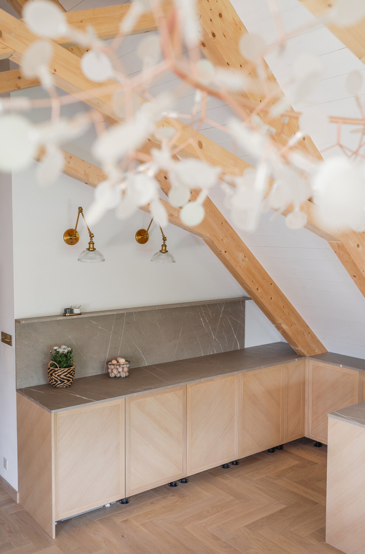 A modern kitchen area features light wooden cabinetry with a smooth stone countertop. Two brass wall sconces are mounted above, providing soft illumination. Exposed wooden beams create a warm atmosphere, and decorative elements are present, including a small potted plant and a stylish centerpiece.