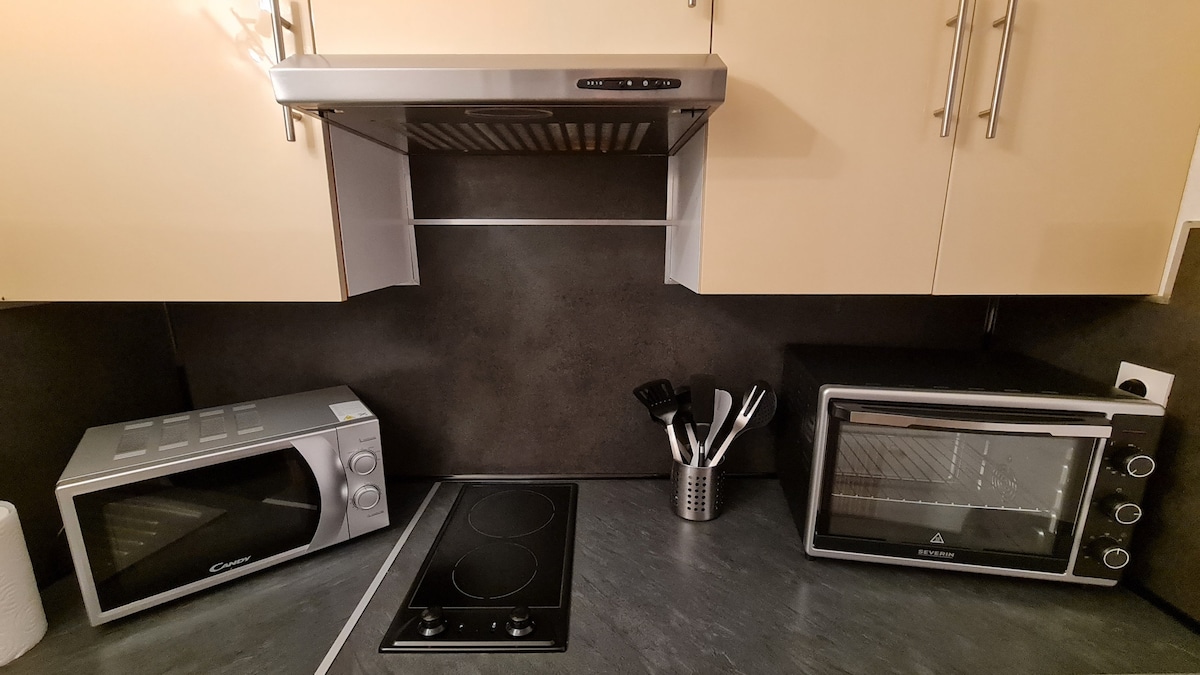 A modern kitchen setup features a compact microwave beside a small oven. A two-burner electric cooktop is integrated into the countertop, while a utensil holder is stocked with various cooking tools. The backsplash offers a sleek, dark surface that complements the cabinetry.