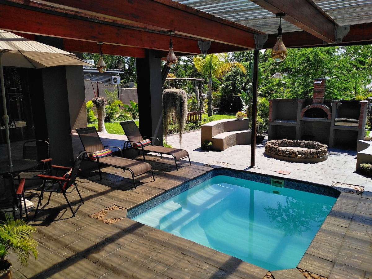 A covered outdoor area features a small swimming pool surrounded by sun loungers. Potted plants and palm trees add greenery, while a built-in barbecue area is visible in the background. Natural light illuminates the space, creating a relaxed atmosphere.