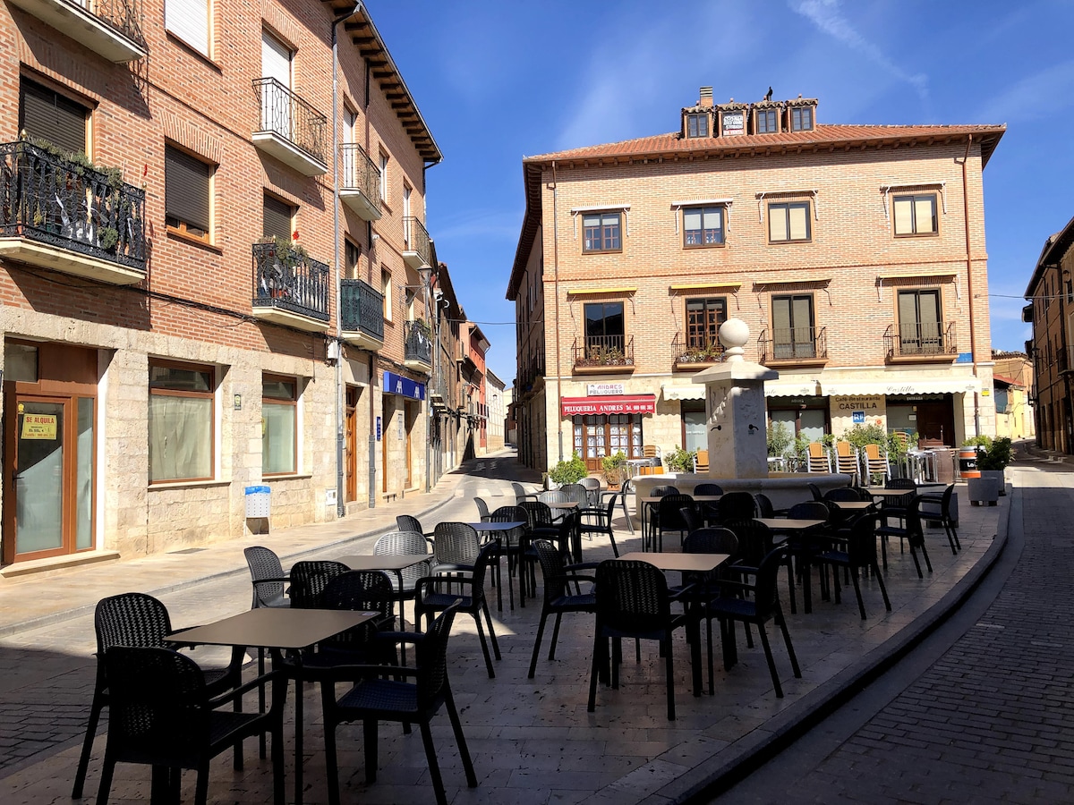 A wide view of a charming town square in Toro features numerous black outdoor chairs arranged around dark tables. The architecture showcases traditional brick buildings with balconies. Sunlight illuminates the scene, highlighting the inviting atmosphere, while a fountain is positioned in the center of the square.