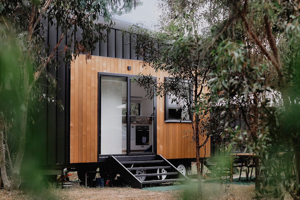The tiny home exterior features a combination of dark metal and warm wooden cladding, framed by lush greenery. Large glass doors open to the outdoor space, providing easy access. A small porch area with steps is visible, surrounded by native bushland.