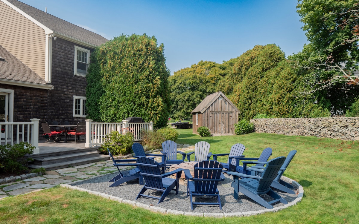 An inviting outdoor space features a circular arrangement of blue Adirondack chairs placed on stone gravel. A wooden table is situated at the center, surrounded by lush green grass and mature trees. A rustic shed can be seen in the background, enhancing the tranquil setting.